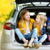 Two adorable girls sitting in a car trunk before going on vacations with their parents. Two kids looking forward for a road trip or travel. Autumn break at school. Family travel by car.