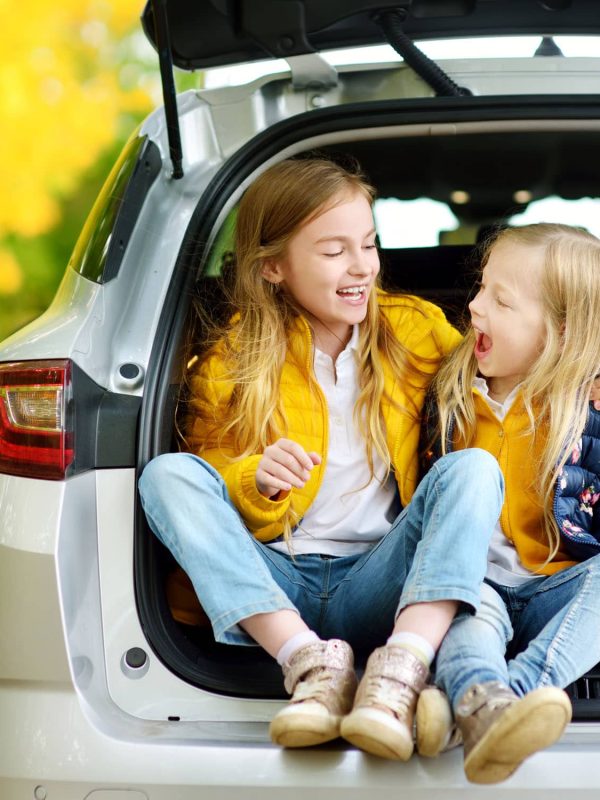Two adorable girls sitting in a car trunk before going on vacations with their parents. Two kids looking forward for a road trip or travel. Autumn break at school. Family travel by car.